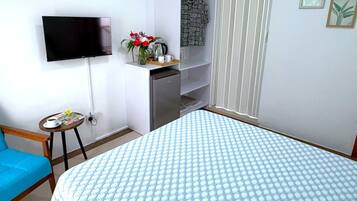 A bedroom with a bed, a TV mounted on the wall, a small table with a cup and magazine, a blue chair, and a kitchenette area.