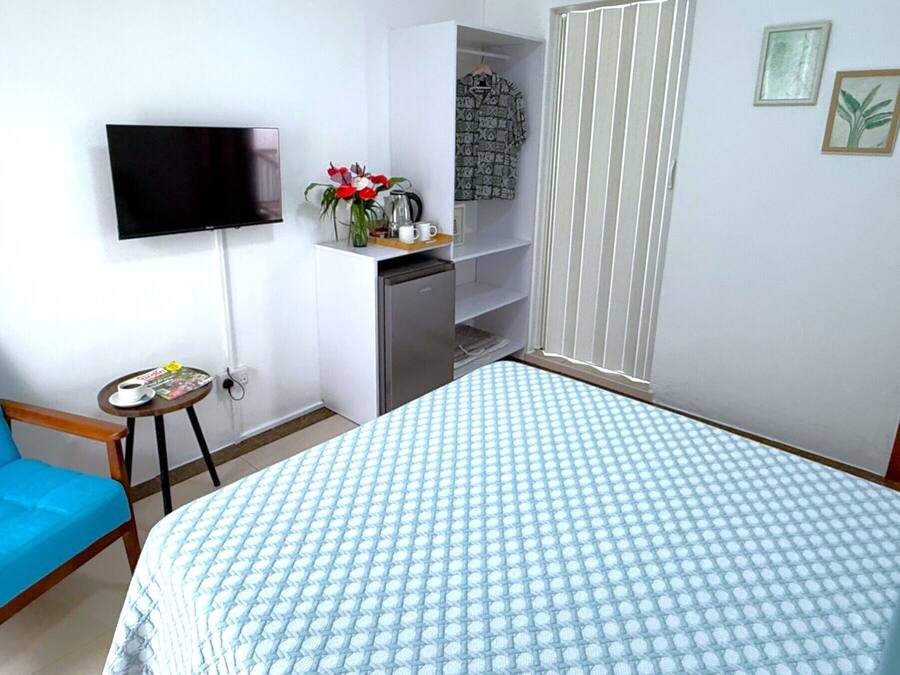 A bedroom with a bed, a TV mounted on the wall, a small table with a cup and magazine, a blue chair, and a kitchenette area.