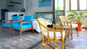 Une chambre d’hôtel moderne avec un lit, deux fauteuils bleus, une chaise en osier, une machine à café, un tableau et une grande fenêtre donnant sur des palmiers.