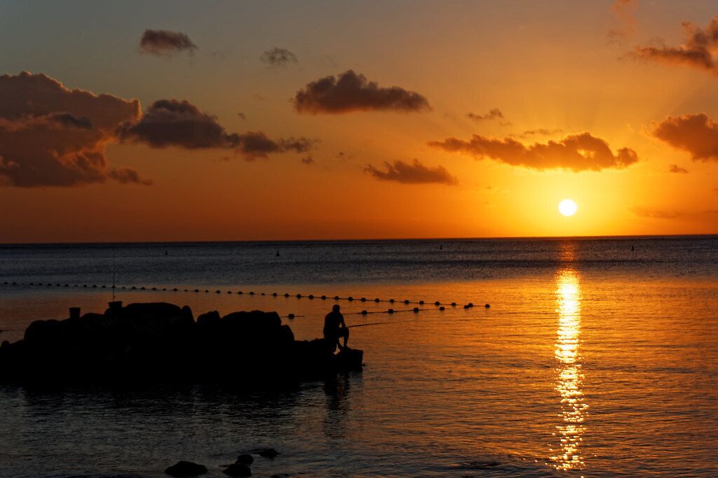 Un atardecer sobre un mar tranquilo, con una persona de pie sobre rocas.