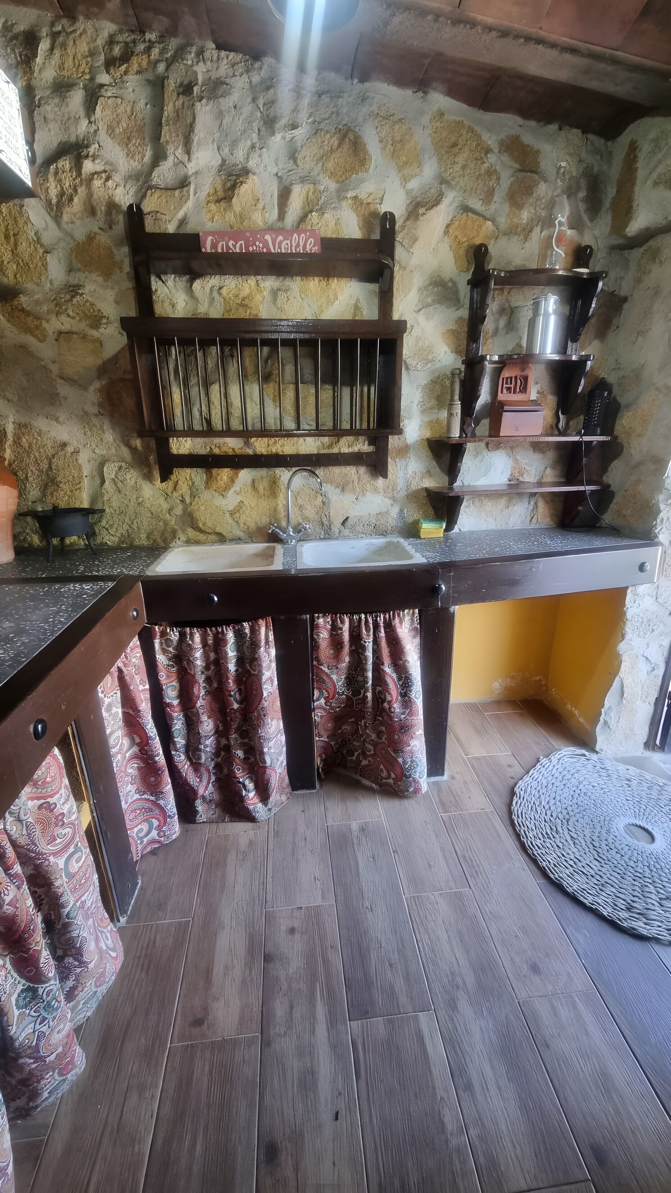 Una cuina rústica amb paret de pedra, armaris de fusta, un rentatall i una barra penjant amb estris de cuina.