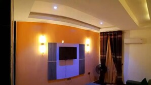 Ein Zimmer mit einem wandmontierten Flachbildfernseher, zwei Wandleuchten, einem Deckenventilator und einem fenster mit Vorhängen.