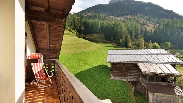 A wooden balcony with a red and white striped chair, a view of a green field, and a rustic building with a metal roof.
