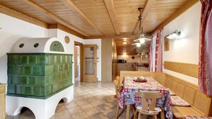 A wooden interior with a dining table, chairs, a bench, a green tiled stove, and a kitchen area.