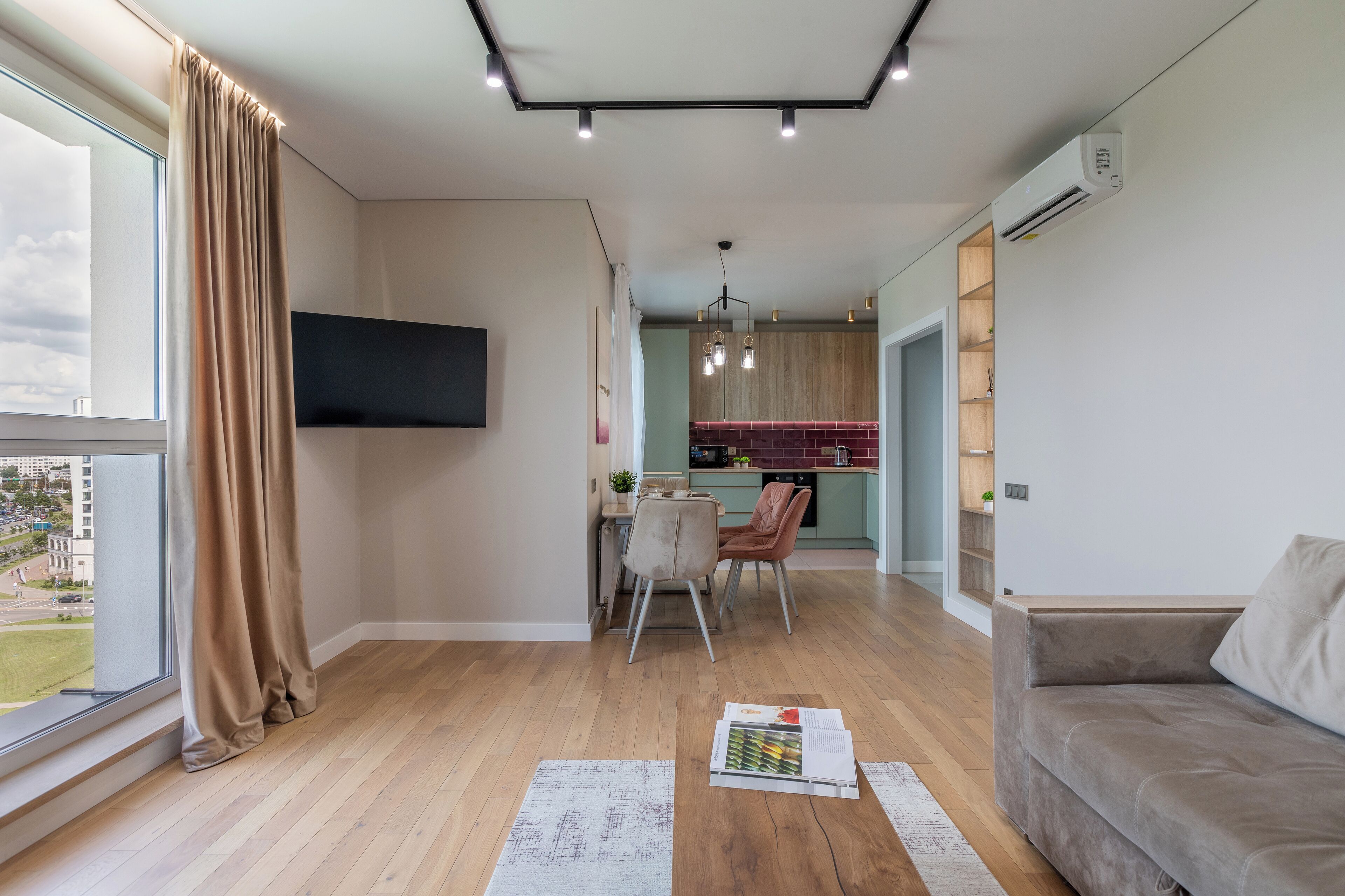 A modern living room with a sofa, a dining area with chairs, a kitchen in the background, and a large window with curtains.