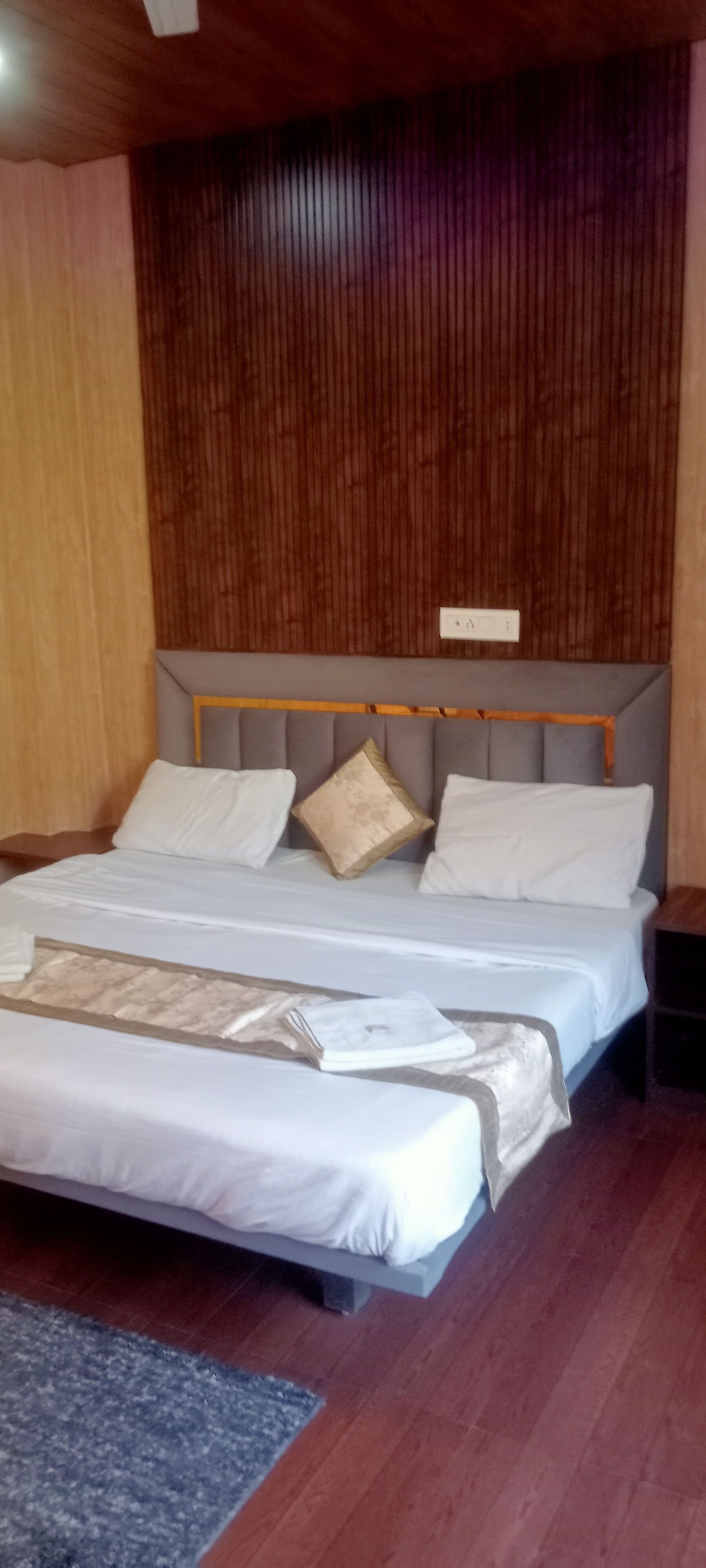 A neatly made bed with white linens and a grey headboard, a wooden floor, and a dark wooden panel wall.