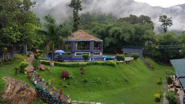 A bungalow with a thatched roof, surrounded by lush greenery and a misty mountain backdrop. - sky ganga resort (Rishikesh)