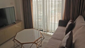 Una sala de estar con un televisor sobre un mueble bajo, una mesa de centro de vidrio, un sofá marrón y cortinas translúcidas.