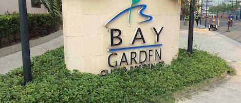 A sign for 'Bay Garden' with a logo, surrounded by greenery and a sidewalk.