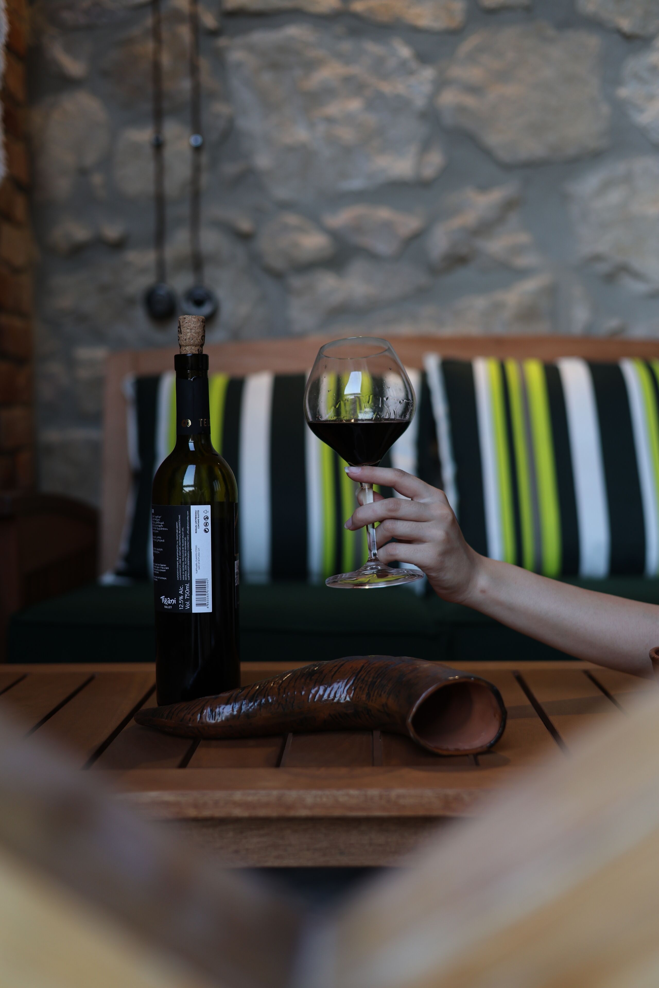 Eine Weinflasche und ein Glas auf einem Holztisch vor einer Steinwand und gestreiftem Kissen