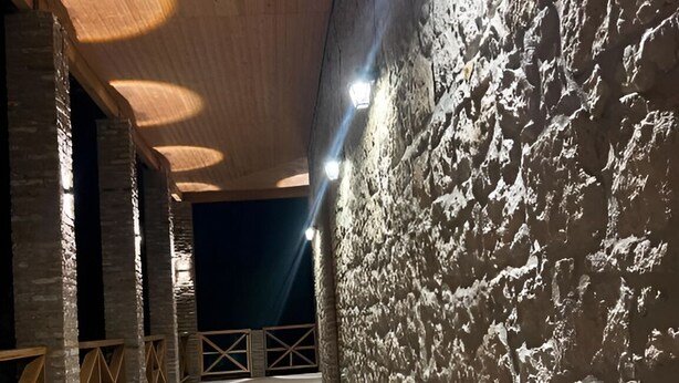 A stone-walled corridor with wooden beams and a concrete floor, illuminated by overhead lights.