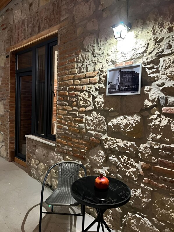 A stone wall with a framed picture, a window with dark shutters, and a metal chair with a table and an apple.