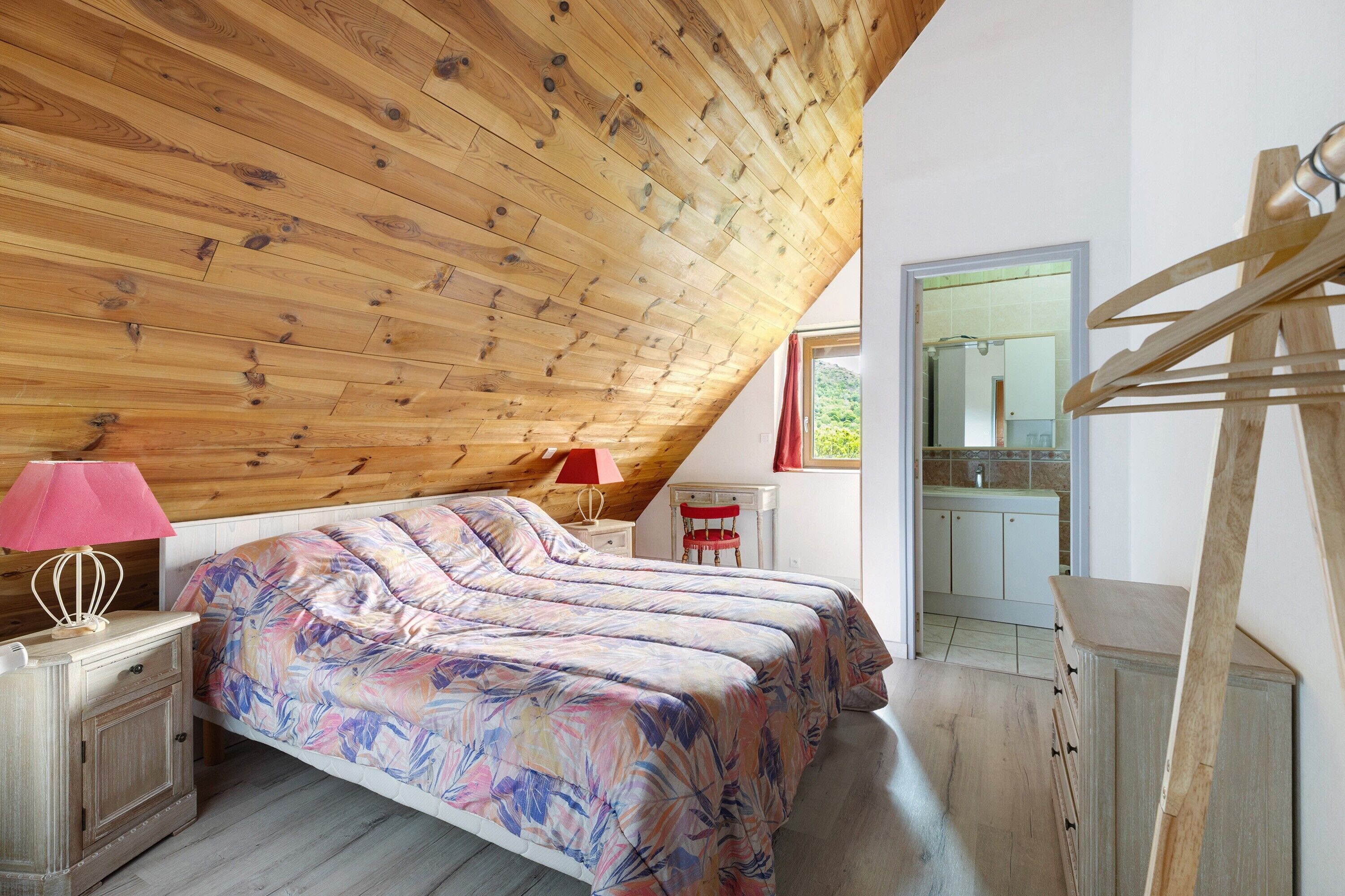 A bedroom with a sloped ceiling, a bed with floral bedding, a nightstand with a lamp, and a view of a kitchen through an open door.