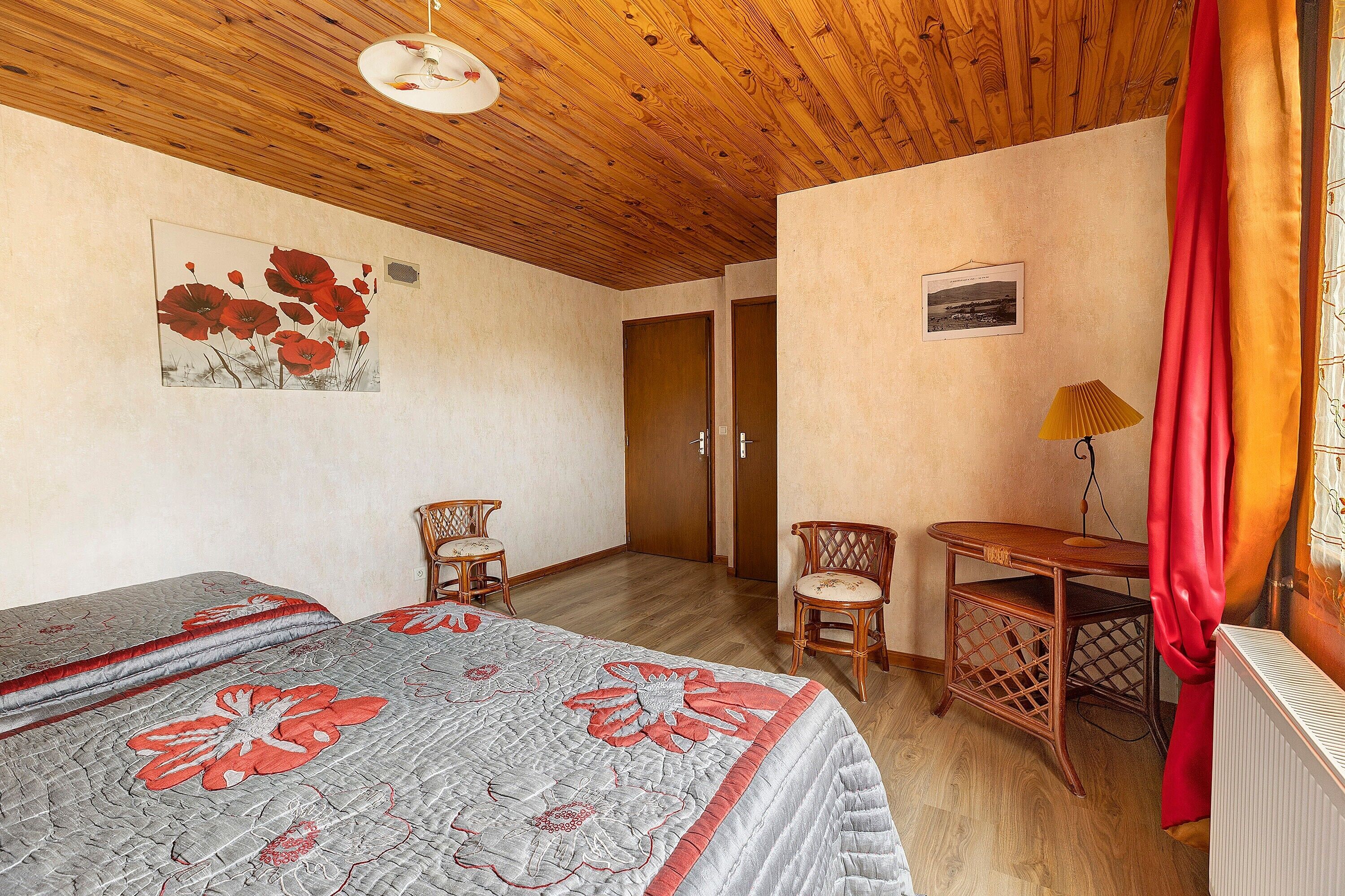 Una habitación con techo de madera, una cama con colcha estampada, dos sillas, una mesita y un radiador.