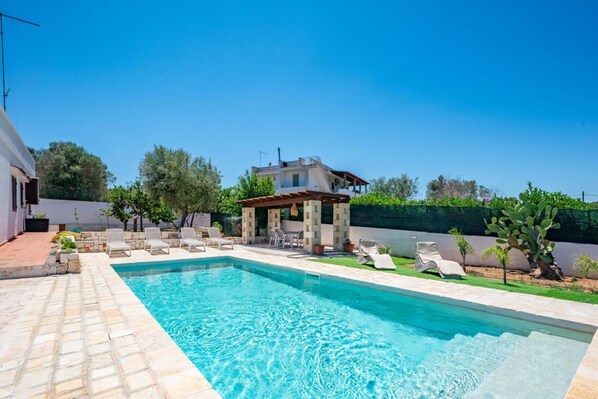 Una piscina con sillas de descanso, una pérgola y una casa al fondo.