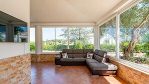 A spacious living area with a large sectional sofa, a brick accent wall, and large windows offering a view of greenery. - Villa Rita Ostuni with pool by Rentbeat  (Ostuni)