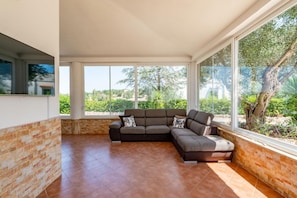 Amplia sala de estar con un sofá modular grande, pared de ladrillo decorativa y ventanas amplias que ofrecen vistas a la vegetación.