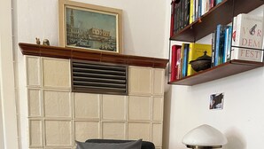 Ein Holzstuhl mit grauem Kissen, ein kleiner weißer Tisch mit Lampe und ein Bücherregal mit Büchern.