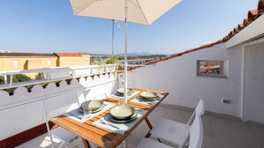 Ein Balkon mit Tisch für vier Personen, einem weißen Sonnenschirm und Blick auf Stadt und Berge im Hintergrund.