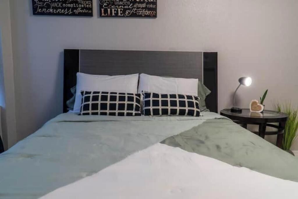 Una cama bien tendida con un cojín a cuadros y una manta verde, una mesita de noche con una lámpara, y decoración de pared con mensajes inspiradores.