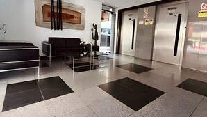 A modern lobby with a reception desk, a black sofa, a glass coffee table, and a large abstract painting on the wall. - Edifício Santorini (Maceio)