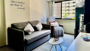 A modern living room with a grey sofa, a chair with a blanket, a small round table with a glass container, and a Heineken fridge. - Edifício Santorini (Maceio)