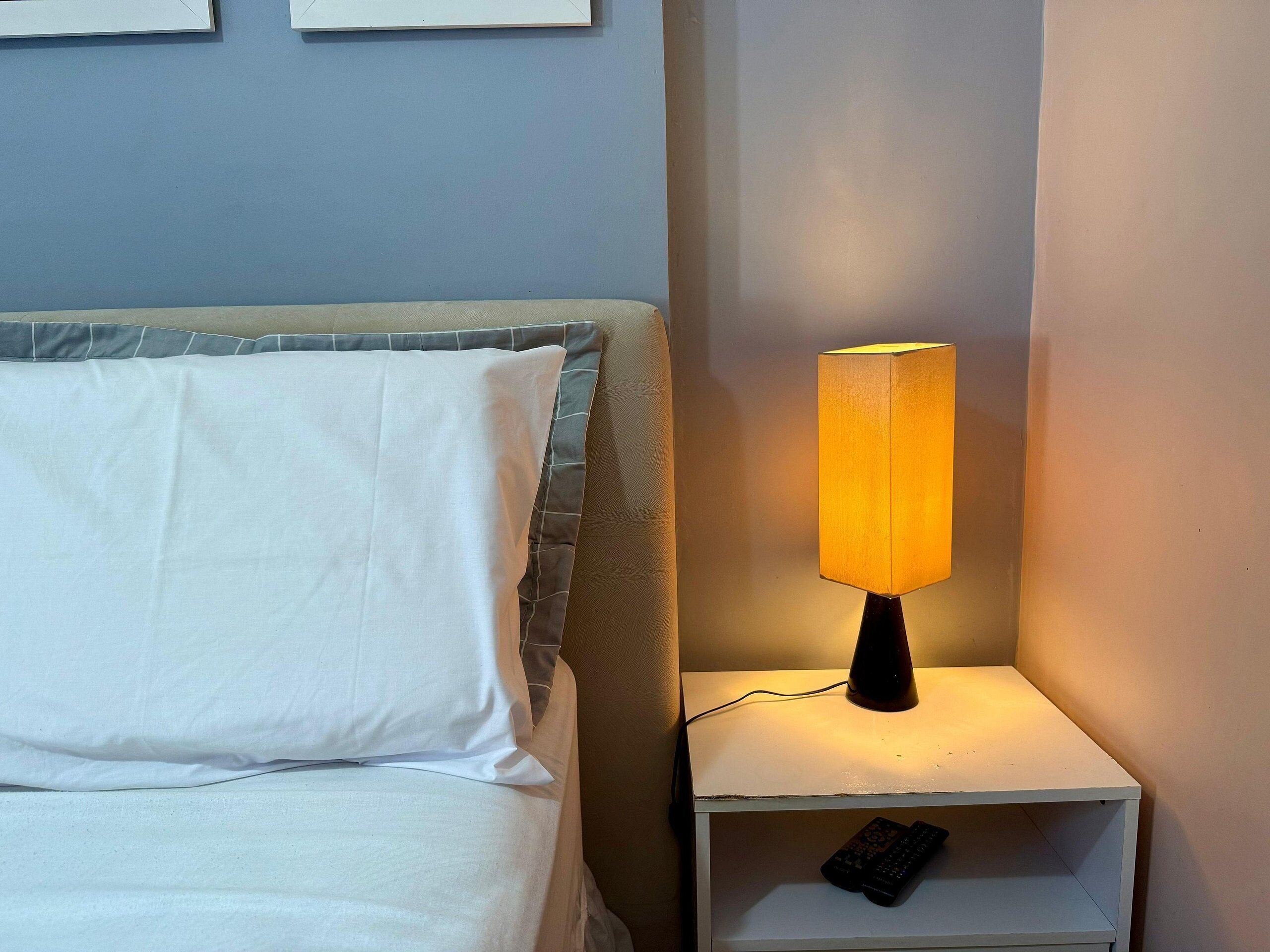 Un lit avec du linge de lit blanc et un oreiller gris, une table de chevet avec une lampe et une télécommande posée sur la table.