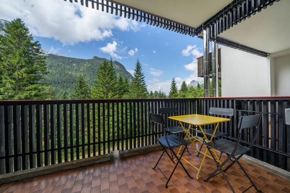 A balcony with a table and chairs, a view of a forested mountain range, and a building with a balcony railing.