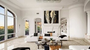 Un salon spacieux avec un canapé blanc, une cheminée, un grand tableau et une vue sur l’extérieur.