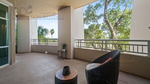 A balcony with a wicker chair, a small table with a plant, and a view of trees and a palm.