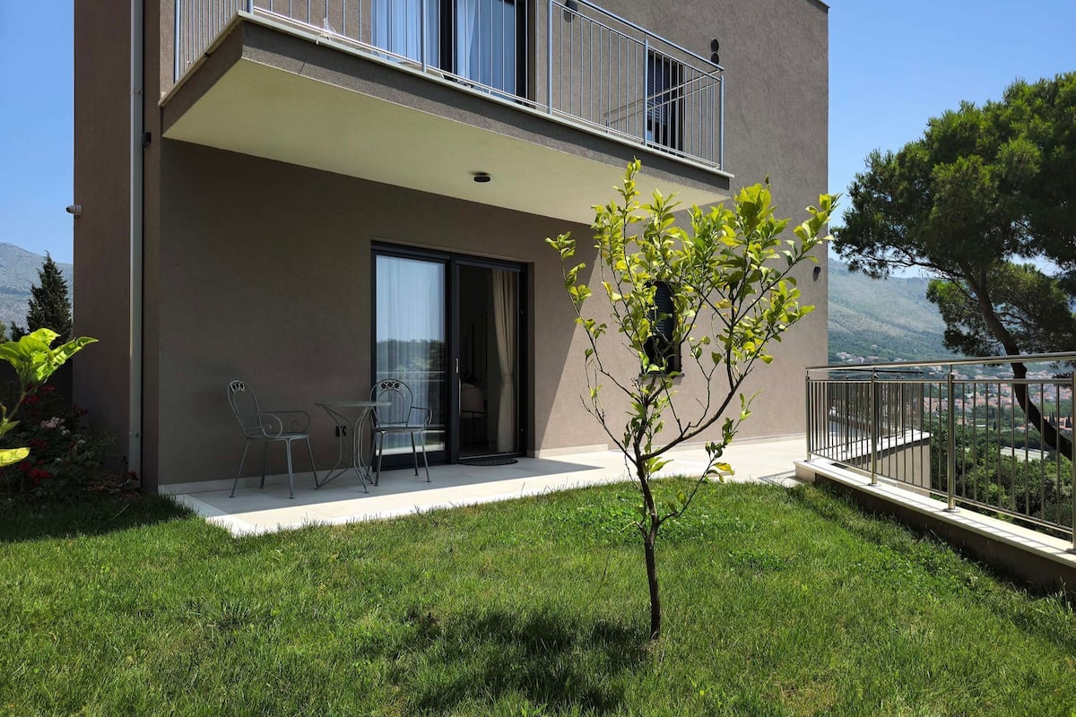 Un edificio moderno de dos pisos con un balcón, un pequeño patio con mobiliario exterior y una zona verde con un árbol joven.