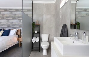 Ein modernes Schlafzimmer mit einem Bett, einem Nachttisch und einem Beistelltisch. Das Badezimmer verfügt über eine Toilette, ein Waschbecken und einen Spiegel.