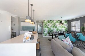 A modern kitchen with a central island, dining area with chairs, and a living space with a sofa and cushions. - Bloubergstrand Apartment, Ocean View & Pool (Cape Town)