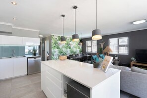 A modern kitchen with white cabinetry, a central island, pendant lighting, and a dining area with a view of the outdoors.