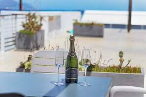 A bottle of Cuvée Brut sparkling wine on a table with two glasses, set outdoors with a view of the sea.