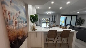 A modern kitchen with a central island, two bar stools, and a large window providing a view of the outdoors. - Let the sun melt your worries away as you unwind in "Searenity" at Riverside (Marco Island)