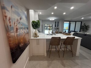 A modern kitchen with a central island, two bar stools, and a large window providing a view of the outdoors. - Let the sun melt your worries away as you unwind in "Searenity" at Riverside (Marco Island)
