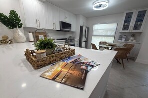 A modern kitchen with white cabinetry, stainless steel appliances, a central island with magazines, and a dining area with wicker chairs.