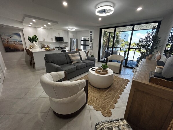 Una sala de estar moderna con un sofá, un sillón y una mesa de centro, conectada a una cocina con armarios blancos y un comedor.