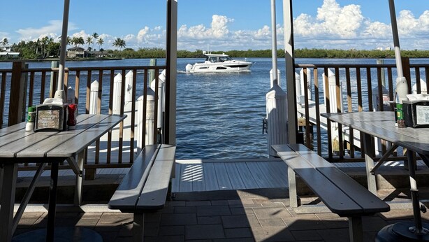 A waterfront patio with tables and umbrellas, overlooking a body of water with a boat. - Let the sun melt your worries away as you unwind in "Searenity" at Riverside (Marco Island)