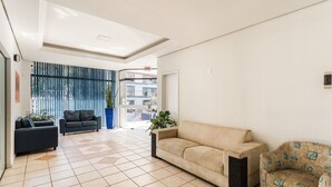 Ampla sala de estar com um sofá bege, um sofá preto e uma poltrona estampada. Há uma grande janela com cortinas azuis e vista para edifícios.