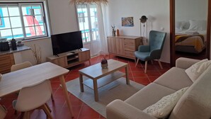 Un salon moderne avec un téléviseur, un canapé, une table à manger et des chaises. On aperçoit en arrière-plan un meuble en bois, un luminaire suspendu et un lit.
