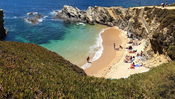 Une plage aux eaux cristallines, bordée de falaises rocheuses et dotée d’une zone de sable où les gens se prélassent au soleil.