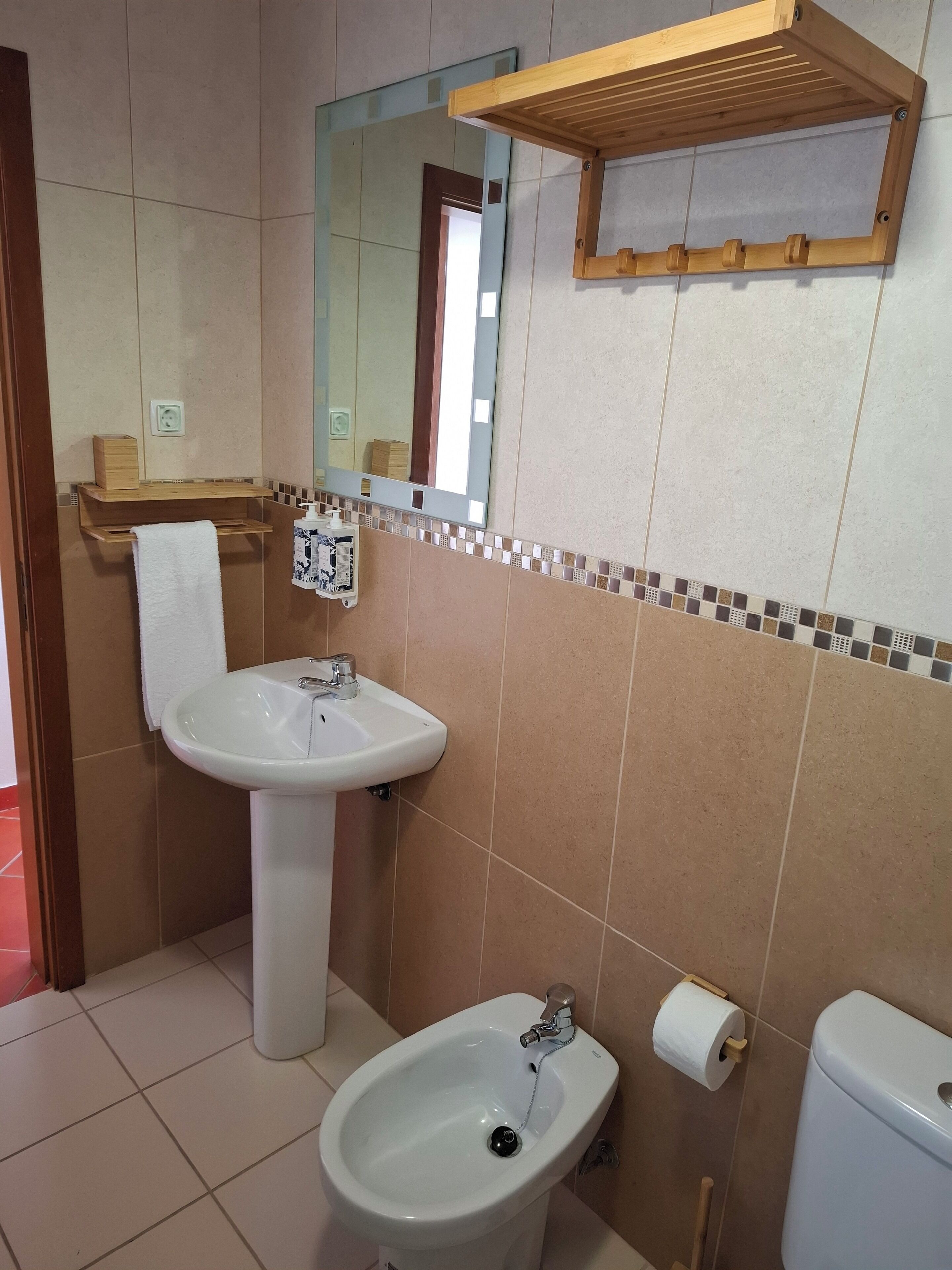 Een badkamer met een wastafel, bidet, toilet en een spiegel met een houten plank erboven.