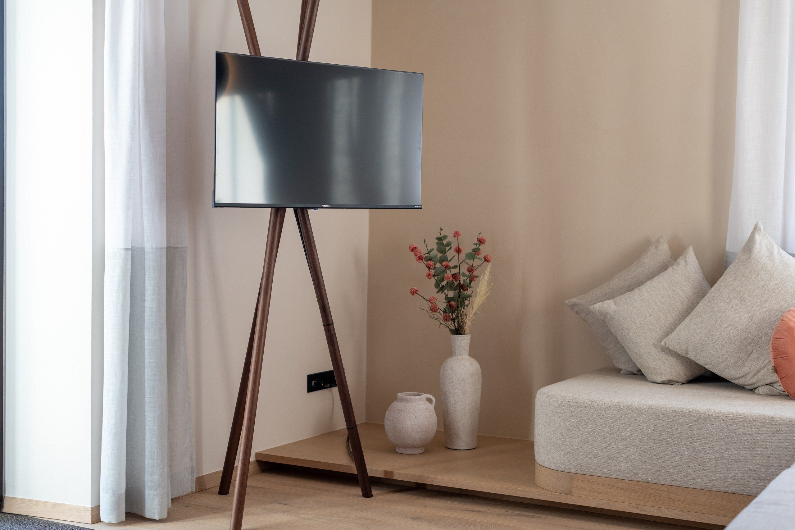 Una habitación minimalista con una televisión de pantalla plana sobre un mueble, un florero blanco con flores y un sofá con cojines.