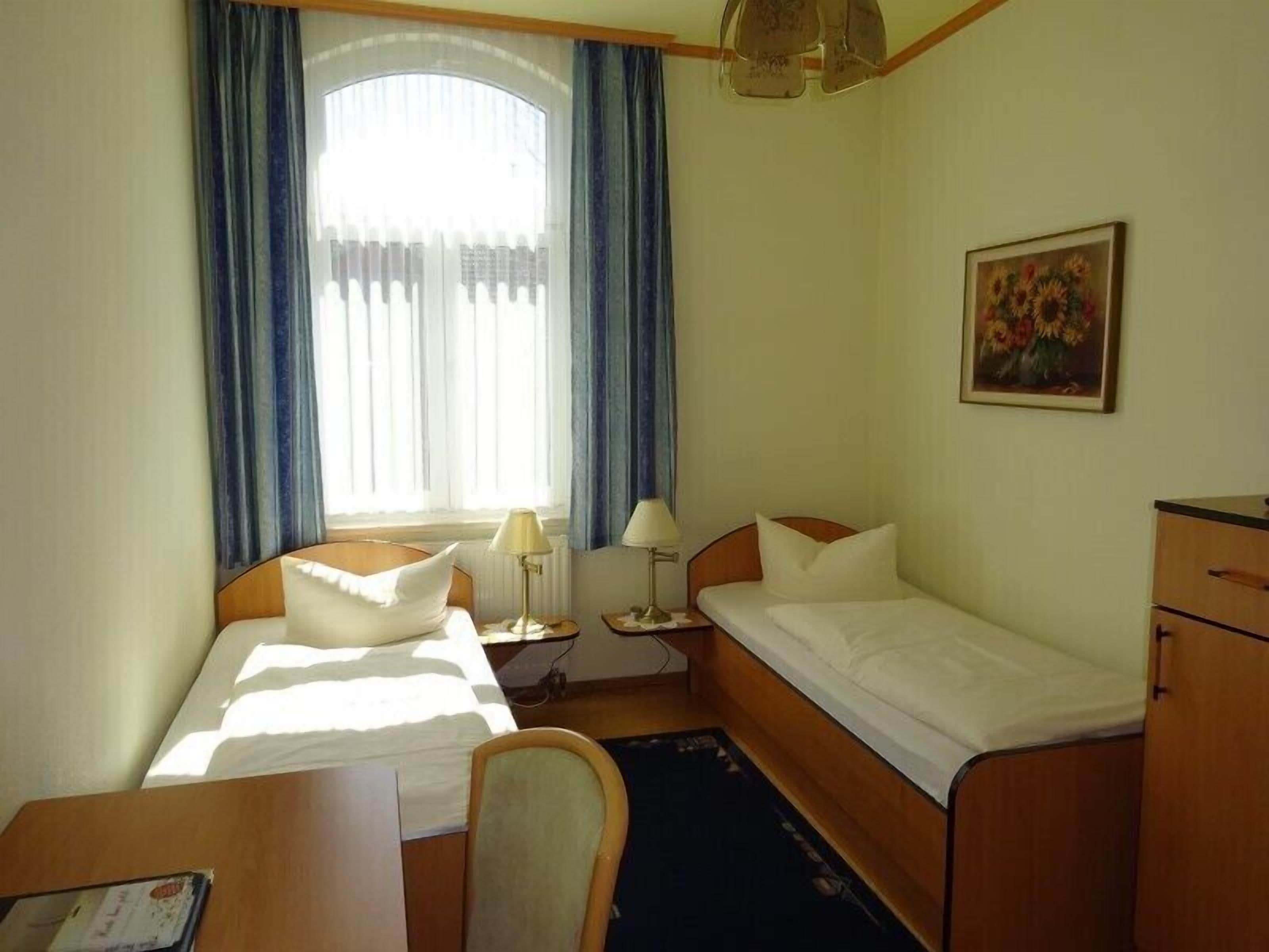 Une chambre d’hôtel avec un lit simple, un bureau, une chaise, une table de chevet, une lampe, une commode et un tableau au mur.