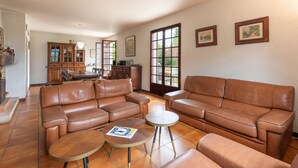 A living room with brown leather sofas, a dining table, and a kitchen in the background. - Villa Delb – Mountain View, Private Pool, and Wi-Fi (Arthez-d'Asson)