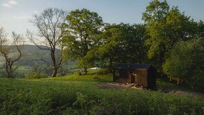 A rustic wooden cabin in a lush green field with a picnic table and umbrella, surrounded by trees and hills. - Off-Grid Woodland Retreat & Wild Sauna Near Llangollen (Llangollen)