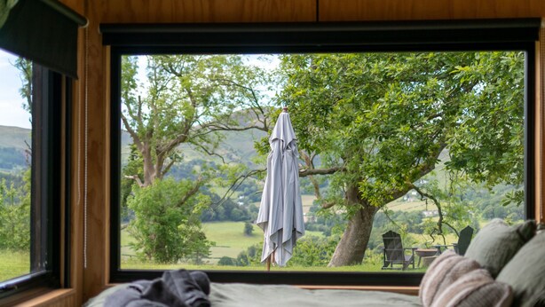 A bedroom with a large window, a bed, and a view of a tree and landscape. - Off-Grid Woodland Retreat & Wild Sauna Near Llangollen (Llangollen)