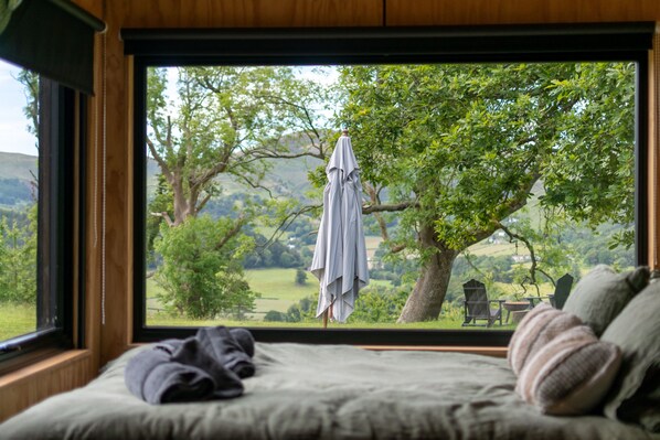 A bedroom with a large window, a bed, and a view of a tree and landscape.